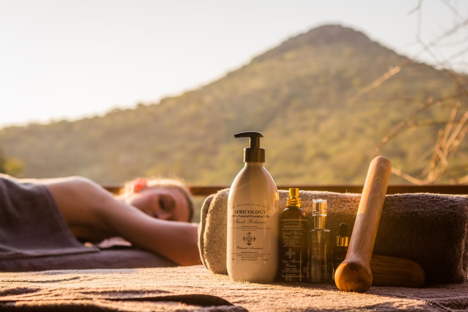 Natural Africology spa products and a wooden massage tool in the foreground, with a guest enjoying an outdoor Safari Recovery Massage in the Zululand bush.